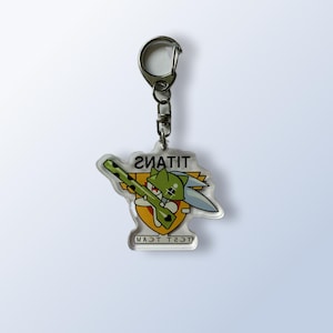 May include: Clear acrylic keychain featuring a green cartoon character with a sword and a rifle. The keychain has the word "TITANS" and "TEST TEAM" printed on it. The keychain is attached to a silver metal clasp.