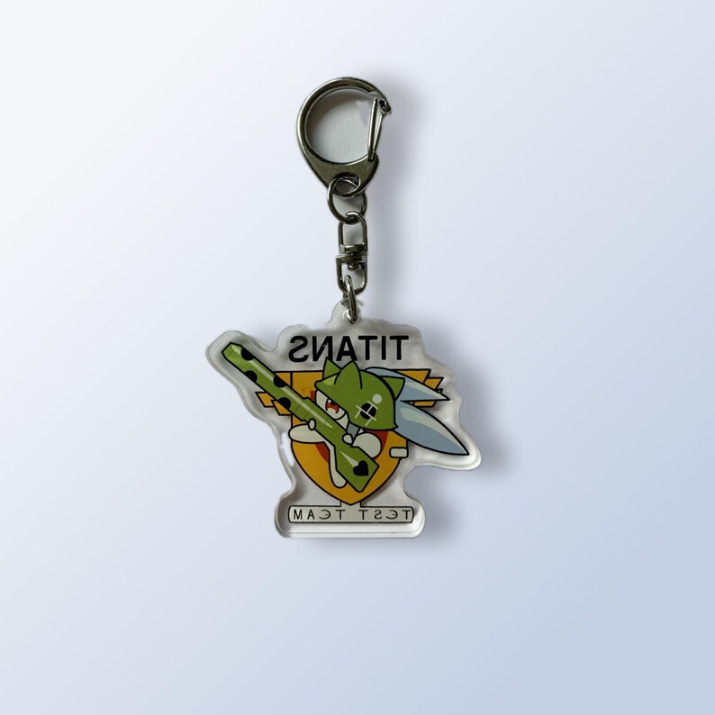 May include: Clear acrylic keychain featuring a green cartoon character with a sword and a rifle. The keychain has the word "TITANS" and "TEST TEAM" printed on it. The keychain is attached to a silver metal clasp.