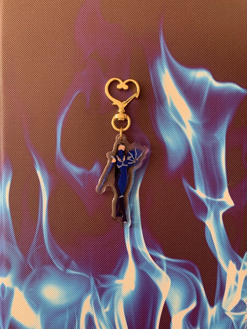 May include: A gold-colored keychain featuring a blue and purple character with wings. The character is standing with arms outstretched and has a heart-shaped ring at the top.