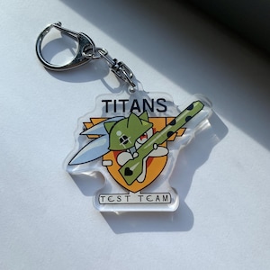 May include: A clear acrylic keychain featuring a cartoon character holding a rifle, with the word "TITANS" above and "TEST TEAM" below. The keychain has a silver metal clasp.