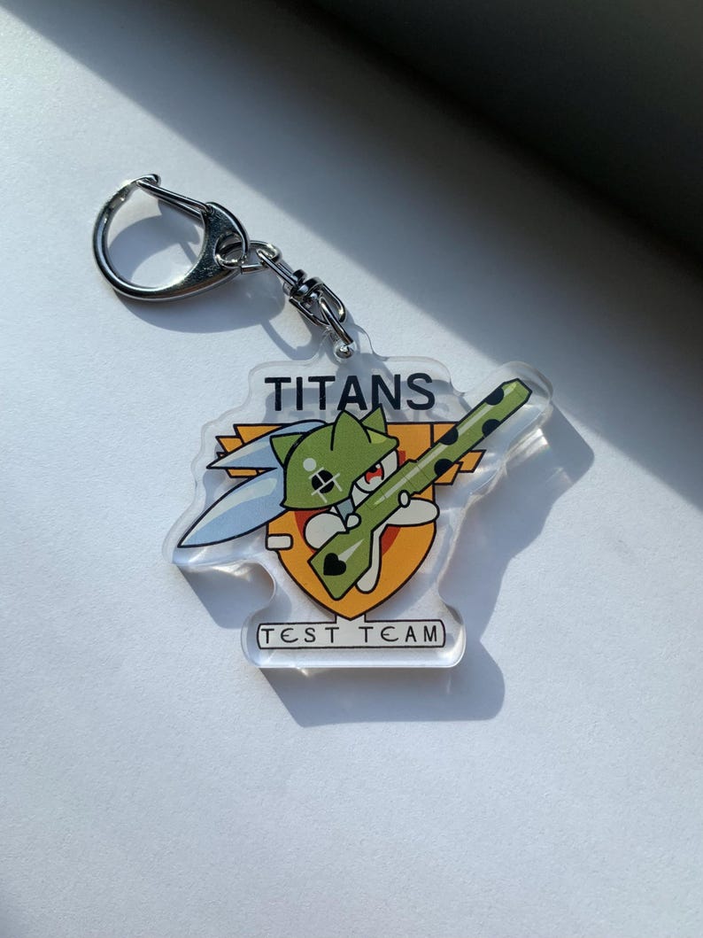 May include: A clear acrylic keychain featuring a cartoon character holding a rifle, with the word "TITANS" above and "TEST TEAM" below. The keychain has a silver metal clasp.