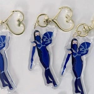 May include: Three blue and white character keychains with gold heart-shaped clasps. Each keychain features a female character in a blue suit with a white cape and a white mask.