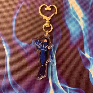May include: A blue and black character keychain with a gold heart-shaped clasp. The character is a female figure with long black hair and a blue outfit.