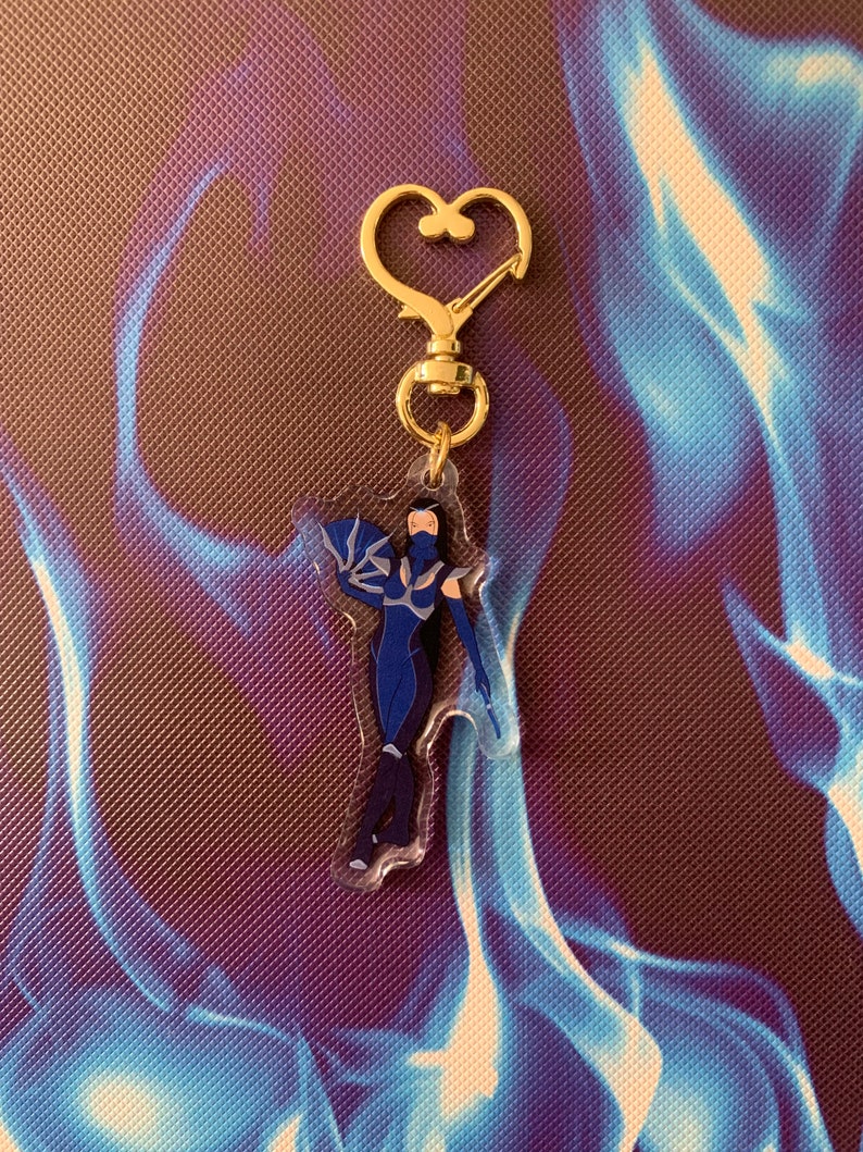 May include: A blue and black character keychain with a gold heart-shaped clasp. The character is a female figure with long black hair and a blue outfit.