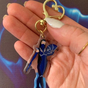May include: A blue and white character keychain with a gold heart-shaped clasp. The character is a woman with a blue mask and a blue outfit. The keychain is made of clear acrylic.