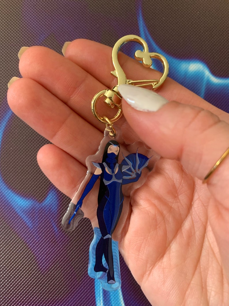 May include: A blue and white character keychain with a gold heart-shaped clasp. The character is a woman with a blue mask and a blue outfit. The keychain is made of clear acrylic.