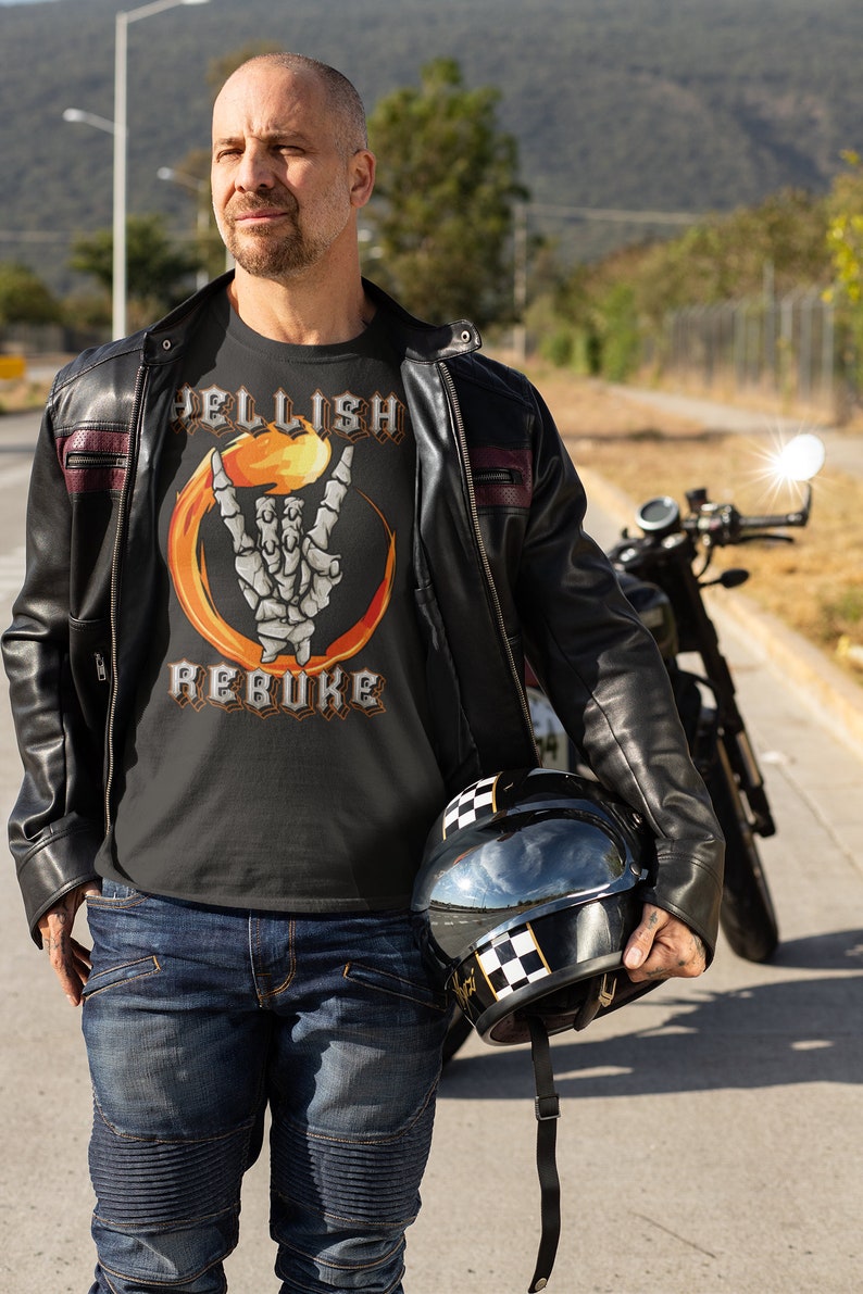 K&ouml;nnte beinhalten: Ein Mann tr&auml;gt ein schwarzes T-Shirt mit einem Grafikdruck einer Skeletthand, die das Rock'n'Roll-Zeichen mit Flammen dahinter macht. Der Text "Hellish Rebuke" ist auf das Shirt gedruckt. Er tr&auml;gt eine schwarze Lederjacke und eine blaue Jeans. Er h&auml;lt einen schwarzen Motorradhelm mit einem karierten Streifen.