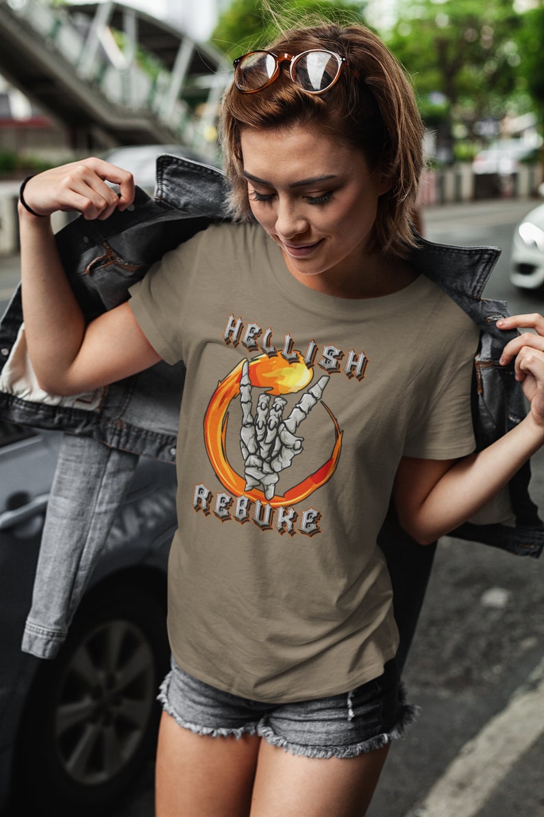 K&ouml;nnte beinhalten: Ein braunes T-Shirt mit einem Grafikdruck einer Skeletthand, die das Rock'n'Roll-Zeichen in einem Feuerkreis macht. Der Text "HELLISH REBUKE" ist in gotischer Schrift um den Kreis herum geschrieben.