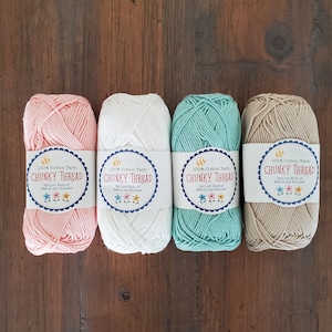 May include: Four skeins of chunky cotton yarn in pastel colors: pink, white, teal, and beige. Each skein has a label that reads "CHUNKY THREAD" and "100% Cotton Yarn" with a bee and flower design. The yarn is arranged on a wooden surface.