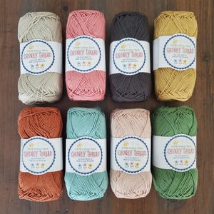 May include: Eight skeins of chunky cotton yarn in a variety of colours, such as beige, pink, grey, and yellow. Each skein has a white label with text and a bee and flower design. The yarn is displayed on a wooden surface.