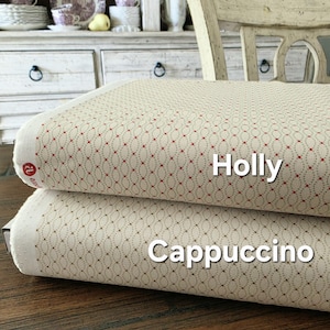 May include: Two fabric samples, labelled "Holly" and "Cappuccino." Both have a cream background with a repeating pattern of red and green dots. The edges have a white border. The fabrics are stacked on a wooden surface, with a blurred background.