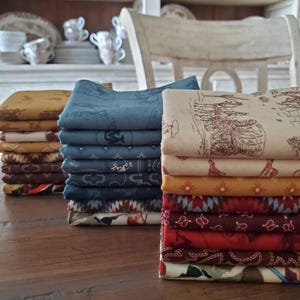 Op de afbeelding: Stapels gevouwen stof in verschillende kleuren en patronen, waaronder blauw, bruin en geel. Sommige stoffen hebben herhalende ontwerpen van paarden, wagens en geometrische vormen. De stoffen zijn gerangschikt op een houten tafel, met een vage achtergrond van een witte stoel en planken.