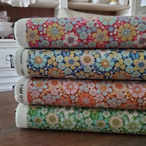 May include: Four rolls of floral fabric in red, navy blue, orange, and green. Each roll displays a repeating pattern of large and small flowers in yellow, pink, and blue. The fabric is labelled with the text "Tilda Quilt Collection".