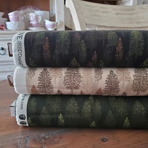 May include: Three rolls of fabric with forest patterns. The top roll is dark with green and brown trees. The middle roll is beige with brown trees. The bottom roll is dark green with green trees. The fabric is stacked on a wooden surface.