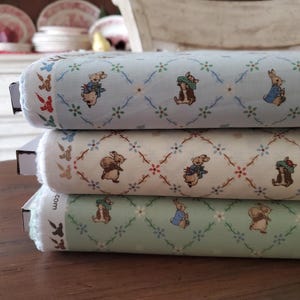 May include: Three fabric rolls stacked, each with a repeating pattern of bunnies in outfits, flowers, and vines. The colours are light blue, cream, and light green. The fabric is likely cotton or a similar material. The rolls are likely for crafting or sewing.