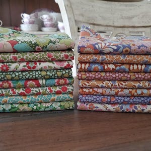 Puede incluir: Dos pilas de tela floral doblada. La pila de la izquierda presenta patrones florales verdes, azules y rojos. La pila de la derecha tiene diseños florales naranjas, morados y rosas. Las telas están apiladas sobre una superficie de madera oscura.