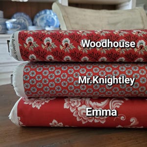 Może przedstawiać: Trzy cylindryczne poduszki dekoracyjne ułożone jedna na drugiej. Górna poduszka jest czerwona we wzór kwiatowy i słowo "Woodhouse". Środkowa poduszka jest czerwona we wzór kołowy i słowa "Mr. Knightley". Dolna poduszka jest czerwona we wzór kwiatowy i słowo "Emma".
