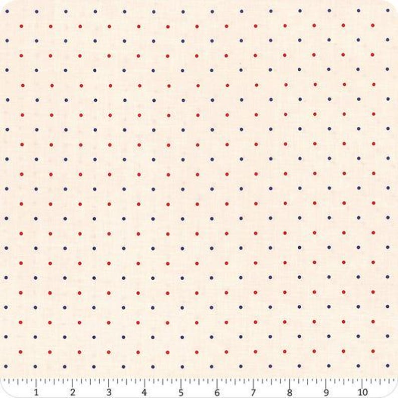 May include: Cream-coloured fabric featuring a repeating pattern of small red and navy blue polka dots. The dots are evenly spaced. A ruler at the bottom shows measurements in inches, equivalent to centimetres.