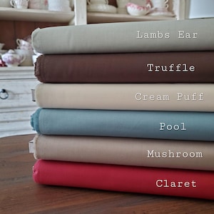 May include: A stack of fabric bolts in various colors, including Lambs Ear, Truffle, Cream Puff, Pool, Mushroom, and Claret. Each bolt is labeled with its color name in white text. The fabrics are neatly stacked on a wooden surface.