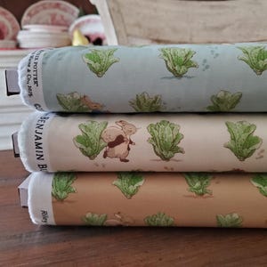 May include: Three rolls of fabric featuring illustrations of rabbits and lettuce. The top roll is light blue, the middle roll is cream, and the bottom roll is tan. The fabric is printed with a repeating pattern of lettuce and rabbits.