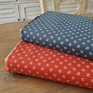 May include: Two folded fabric pieces, one red and one navy blue, with a repeating white floral and dot pattern. The fabrics are stacked on a wooden surface, showcasing the detailed design and texture of the material. The fabrics are likely for sewing or crafting.