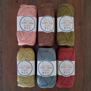 May include: Six skeins of chunky cotton yarn in various colors, including pink, brown, and green. Each skein has a label that reads "100% Cotton Yarn CHUNKY THREAD" with a floral and bee design. The yarn is arranged on a wooden surface.