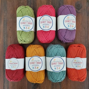 Könnte beinhalten: Draufsicht auf acht Knäuel farbenfrohen, groben Baumwollgarns. Die Farben umfassen Grün, Pink, Lila, Rot, Gelb, Türkis und Orange. Jedes Knäuel hat ein weißes Etikett mit dem Text "CHUNKY THREAD" und "100% Cotton Yarn".