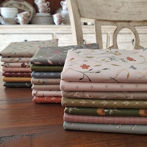 May include: Stacks of floral patterned fabric in various colors, including pink, green, and gray, are arranged on a wooden surface. The fabric features delicate floral and leaf designs. The background includes a white chair and a cabinet with decorative dishware.
