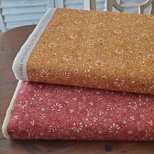 May include: Two rolls of fabric, one in a warm mustard yellow and the other in a deep red. Both fabrics feature a delicate floral pattern with white and red accents. The text "Love Struck" by Kim Diehl for Henry Glass is visible on the edge of the yellow fabric.