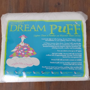 May include: A package of white Quilters Dream Puff batting featuring a blue label with a cartoon goose wearing a hat and a colourful quilt. The text includes product benefits, usage instructions, and a list of available sizes from craft to king.