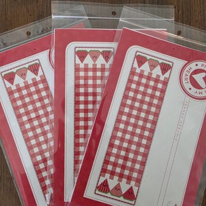 Puede incluir: Tres marcadores de libros en fundas de plástico. Cada marcador presenta un patrón de cuadros rojos y blancos con rodajas de sandía en la parte inferior y superior. Los marcadores están sobre un fondo rojo. El texto "Yum!" y "#848" son visibles.