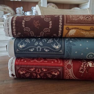 May include: Three rolled bandanas in shades of brown, blue, and red. Each bandana features a unique pattern with paisley designs, horseshoes, and other western-themed motifs. The bandanas are stacked on top of each other.