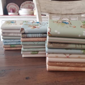 Pode incluir: Pilhas de tecido com vários padrões pastel, incluindo coelhos e desenhos florais. O tecido é em tons de azul, verde e bege, alguns com texto. As pilhas estão dispostas numa mesa de madeira, com uma cadeira branca e pratos decorativos no fundo.