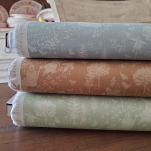 Puede incluir: Tres rollos de tela en colores pastel, apilados. El rollo superior es azul claro, el del medio es marrón claro y el inferior es verde claro. Cada rollo presenta un patrón repetido de árboles, flores y animales.