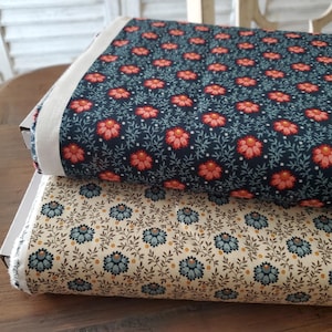 Op de afbeelding: Twee rollen stof met bloempatronen. Eén rol heeft een marineblauwe achtergrond met rode en roze bloemen en groene bladeren. De andere rol heeft een crèmekleurige achtergrond met blauwe bloemen en bruine bladeren. Beide liggen op een houten oppervlak.