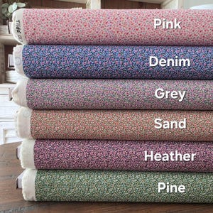 Peut inclure: Une pile de rouleaux de tissu aux couleurs variées : rose, denim, gris, sable, bruyère et pin. Chaque rouleau est orné d'un petit motif floral. Les mots "Pink", "Denim", "Grey", "Sand", "Heather" et "Pine" sont écrits sur le tissu.