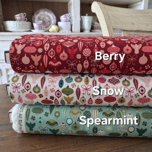 May include: Three rolls of Christmas fabric in Berry, Snow, and Spearmint colours. Each fabric features a repeating pattern of ornaments, bows, and festive shapes. The Berry fabric is a deep red, the Snow fabric is white, and the Spearmint fabric is a light green.
