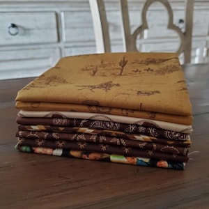 May include: A stack of folded fabric squares with various Western-themed patterns. The top fabric features a desert landscape with cowboys and cacti in shades of brown and tan. Other fabrics have paisley and floral designs.