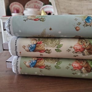 Pode incluir: Três rolos de tecido empilhados. O tecido tem um fundo verde claro, creme e azul claro com um padrão repetido de coelhos, cenouras e outros elementos de jardim. Os textos "Rights Reserved" e "www.RileyBlake" são visíveis.