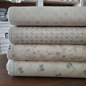 以下が含まれることがあります： さまざまな模様の4つの生地ロールのスタック。一番上のロールは、クリーム色の背景にグレーのボタンのデザインが繰り返されています。2番目のロールは、青とグレーの花柄です。3番目のロールは、茶色とクリーム色の花柄です。一番下のロールは、スプールと針のデザインです。