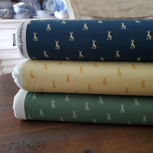 May include: Three rolls of fabric in navy, cream, and green, each patterned with small gold or cream-coloured rabbit silhouettes. The fabric is stacked on a wooden surface, with the edges of the rolls visible.