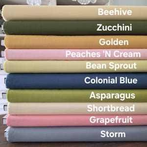 Könnte beinhalten: Ein Stapel gefalteter Stoffe in verschiedenen Farben, darunter Beige, Olivgrün, Gold, Rosa, Hellgrün, Marineblau und Grau. Jeder Stoff ist mit seinem Farbnamen beschriftet, wie "Beehive" und "Zucchini".