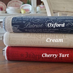 Può includere: Tre rotoli di tessuto nei colori patriottici: blu navy con testo bianco, crema con testo beige e rosso con testo bianco. Il rotolo blu navy è etichettato "Oxford Patriot", il rotolo crema "Cream Independence" e il rosso "Cherry Tart".