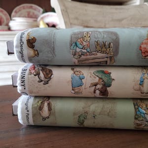 Puede incluir: Tres rollos de tela con ilustraciones de conejos en varias escenas. La tela tiene un fondo azul claro, crema y verde. El texto "BlakeDesigns.com" y "BUNNY" son visibles en los bordes. Las ilustraciones representan escenas de un cuento infantil.