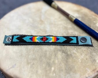 Könnte beinhalten: Ein Armband mit Perlen mit türkisfarbenem Hintergrund und einem zentralen Design aus roten, gelben und blauen Perlen. Das Armband hat schwarze Perlenakzente und einen schwarzen Druckknopfverschluss. Eine Trommel und ein Trommelstock befinden sich im Hintergrund.