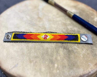 Könnte beinhalten: Ein Lederarmband mit einem bunten geometrischen Muster aus roten, gelben, blauen und weißen Perlen. Das Armband hat einen silbernen Verschluss.