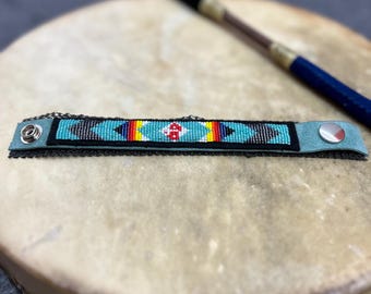Könnte beinhalten: Ein türkisfarbenes Armband mit Perlenbesatz und einem geometrischen Muster in Rot, Gelb, Schwarz und Weiß. Das Armband liegt auf einer Trommel, im Hintergrund ein Trommelstock. Das Armband hat einen silbernen Knopfverschluss und eine schwarze Spitzenborte.
