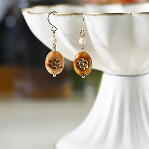 May include: A pair of gold dangle earrings with brown and white oval beads and a gold flower design. The earrings are hanging from a white pedestal.