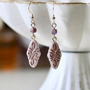 May include: A pair of gold earrings with a light pink oval-shaped bead with a brown design. The beads are attached to a gold chain with a small purple bead.
