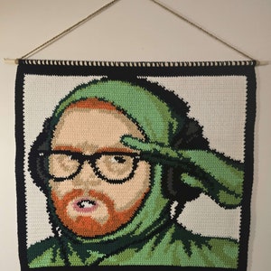 Puede incluir: Un tapiz de pared tejido a ganchillo con un retrato de una persona con gafas, barba roja y capucha verde. La obra de arte está enmarcada con un borde negro y colgada con una barra de madera y una cuerda.